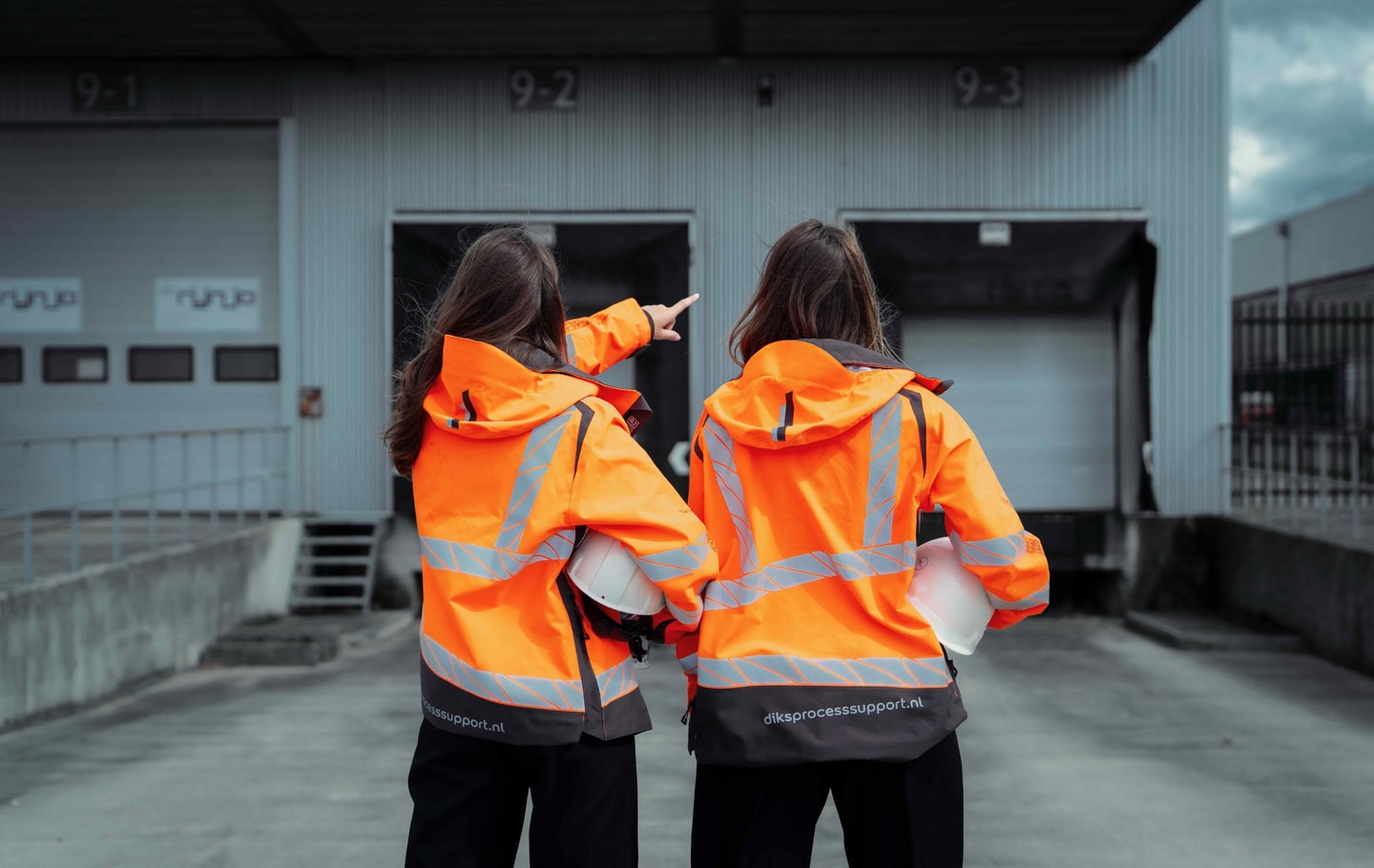 Twee vrouwen in oranje veiligheidsjassen wijzen naar de laaddocks van een distributiecentrum