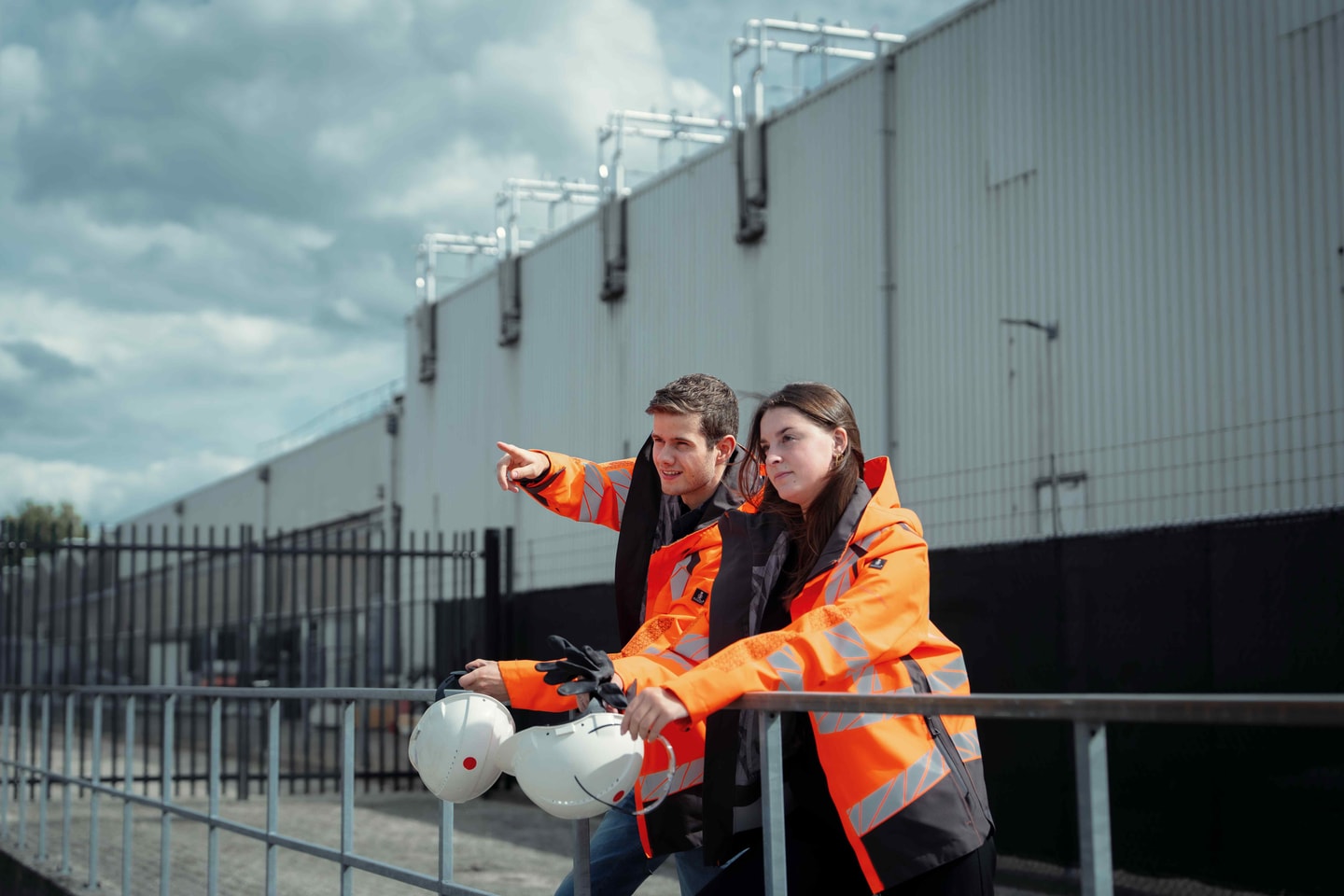 Twee mensen in oranje jassen met helmen bij een hek voor een bedrijfshal
