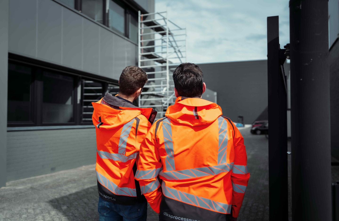 Twee mannen in oranje jassen kijken omhoog naar een steiger naast een gebouw