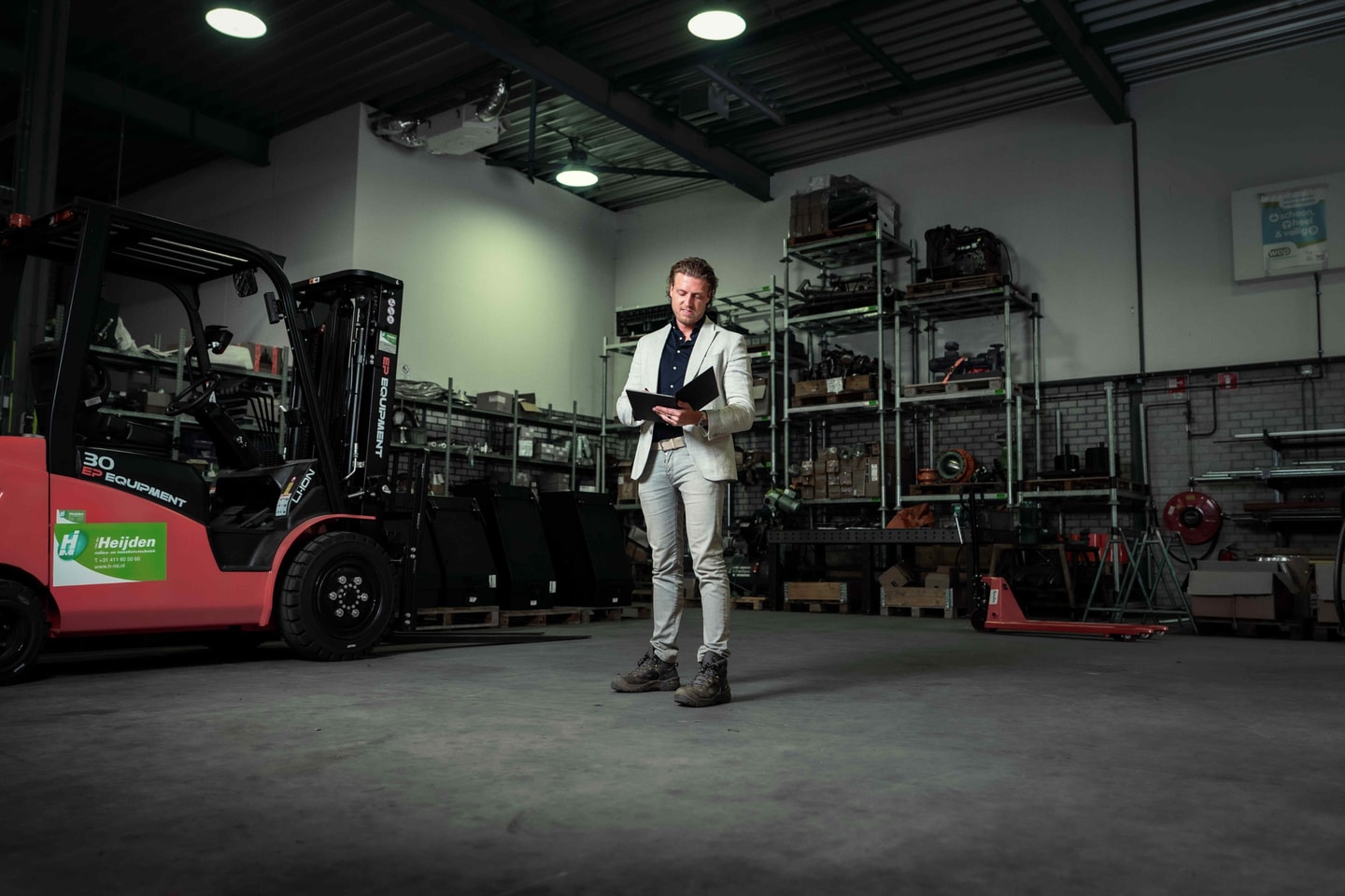 Man in colbert staat met een tablet in een magazijn met een rode heftruck