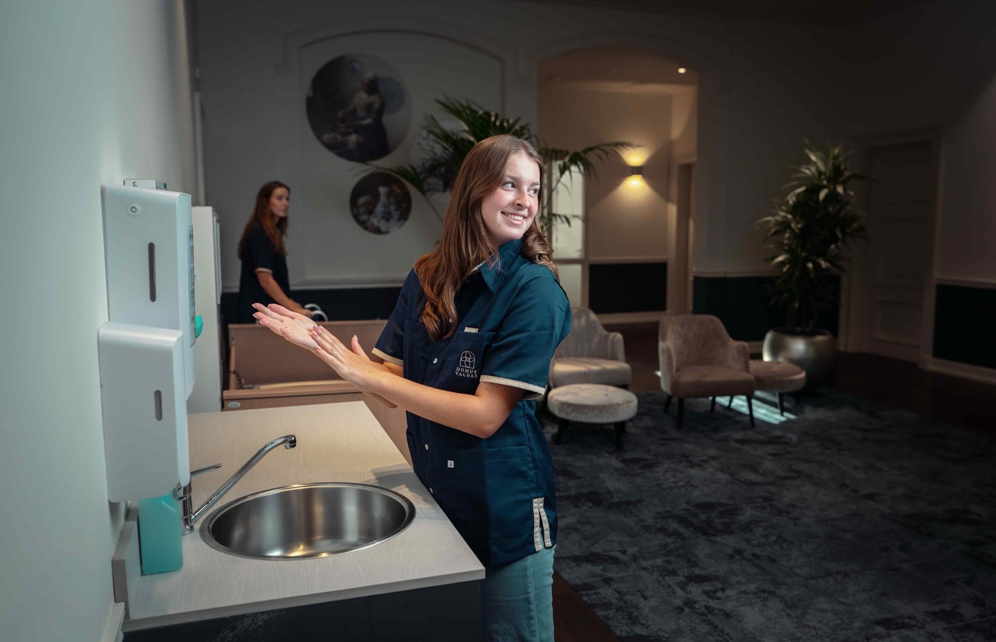 Medewerker in zorguniform bij een wasbak in een ontvangstruimte met lounge