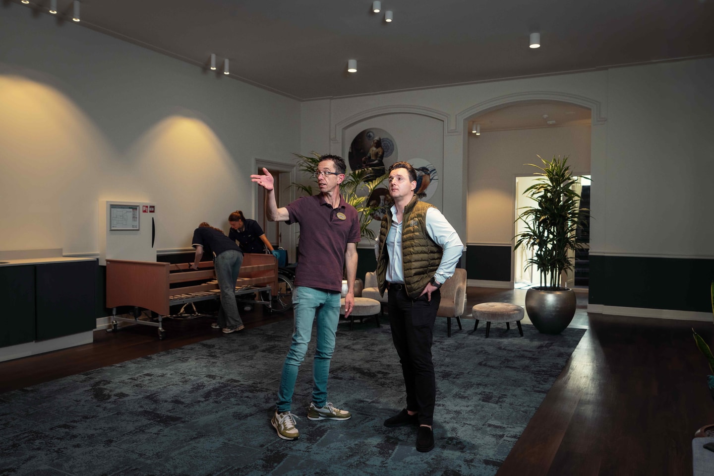 Twee mannen in gesprek in een ruime hal met wandlampen en een plant