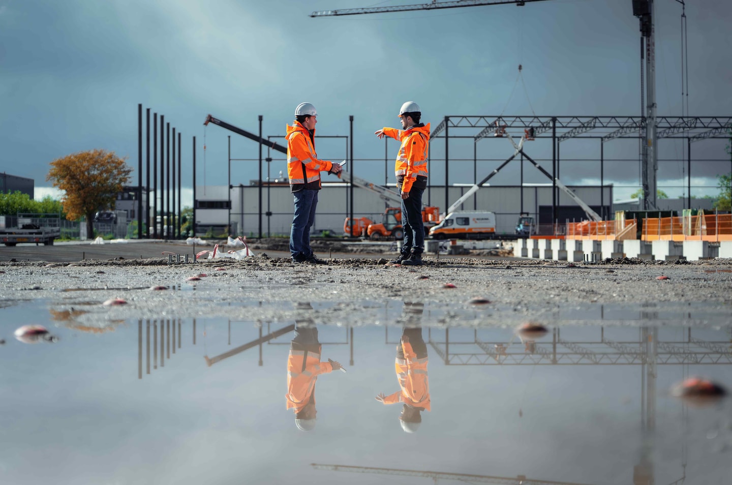Twee mensen in oranje werkkleding op een bouwterrein dat weerspiegelt in een plas