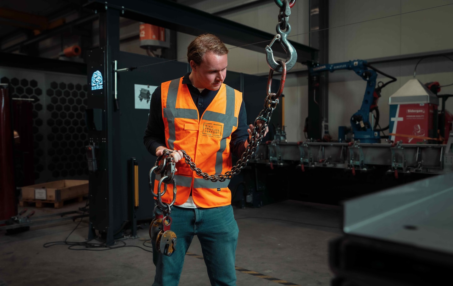 Man in oranje hesje in een fabriekshal, met een hijsketting naast hem