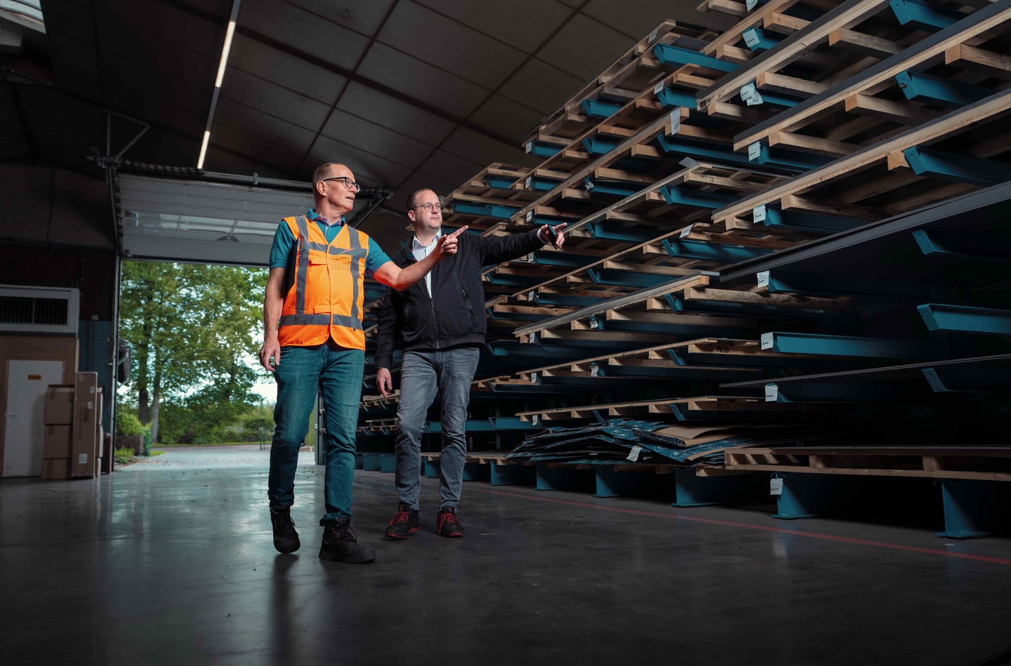 Twee mannen, een in oranje hesje, in gesprek in een loods met lange stellingen