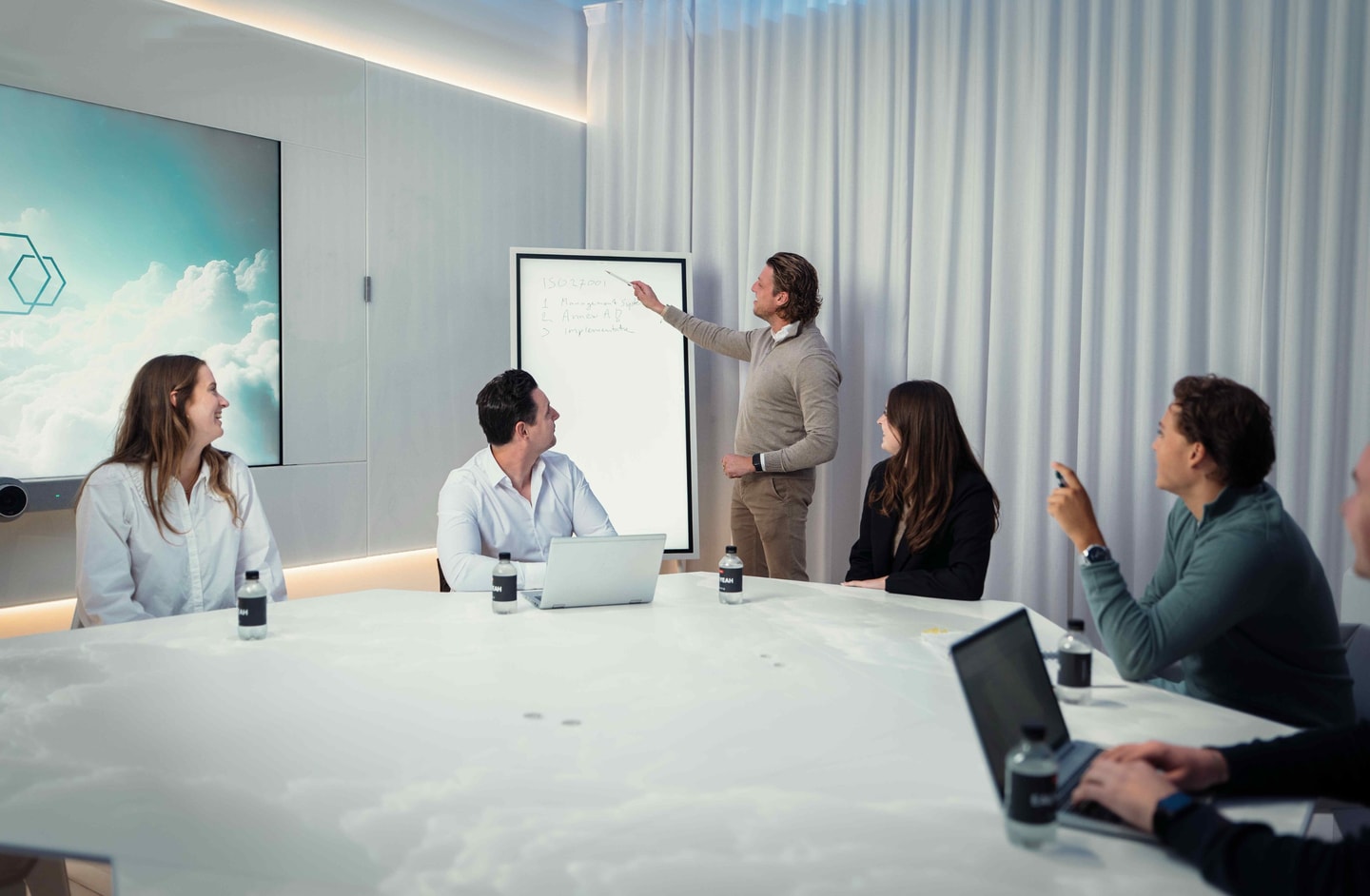 Man presenteert aan een digitaal scherm voor vier collega’s aan een witte vergadertafel