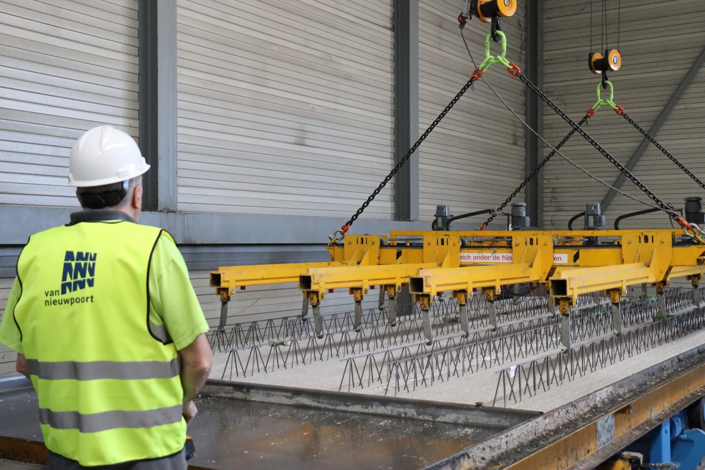 Medewerker met helm en geel hesje kijkt naar een hijsbalk boven een betonbekisting in een fabriekshal
