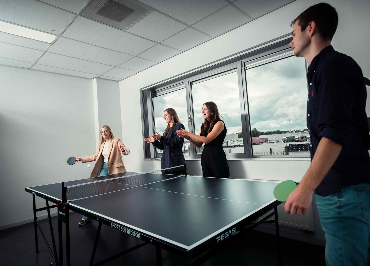 Collega’s aan een tafeltennistafel bij een raam met uitzicht op een bedrijventerrein